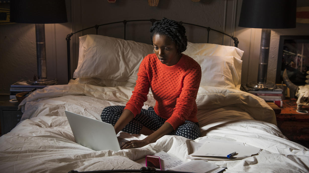  A woman sits on a bed in a dark room, typing on a laptop. 