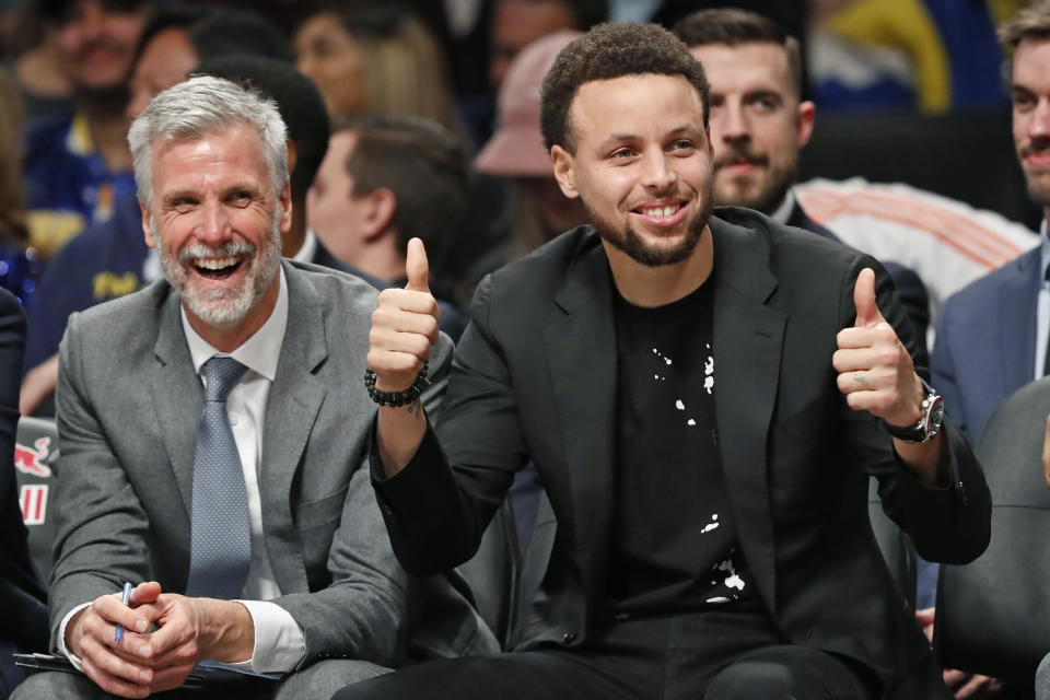 ADDS NAME OF ASSISTANT COACH - Golden State Warriors Stephen Curry, right, gives a thumbs-up to a Brooklyn Nets fan known as "Mr. Whammy" during the second half of an NBA basketball game Wednesday, Feb. 5, 2020, in New York. At left is Warriors assistant coach Bruce Fraser. Curry is recovering from a hand injury. (AP Photo/Kathy Willens)