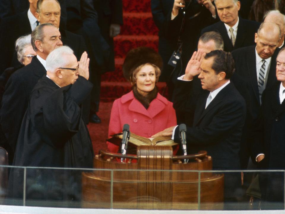 Pat in a bright pink coat and fur scarf and hat at a podium next to her Richard Nixon.
