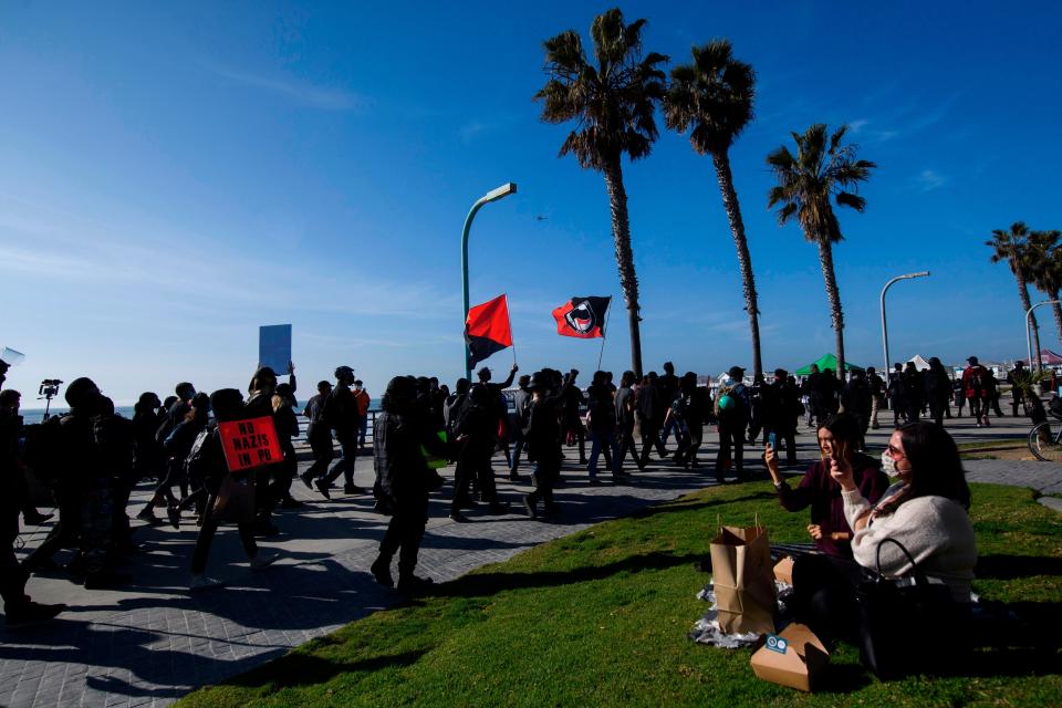 People eating a meal at the beach take pictures as counter-protesters, some carrying Antifa flags, march in opposition to demonstrators holding a "Patriot March" in support of President Donald Trump on January 9, 2021 in the Pacific Beach neighborhood of San Diego, California.