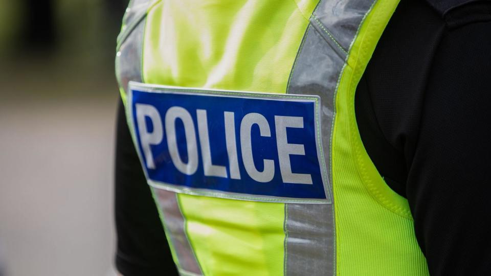 A police officers back, they are wearing a black top with a high vis vest over the top which has a badge reading Police on a blue background