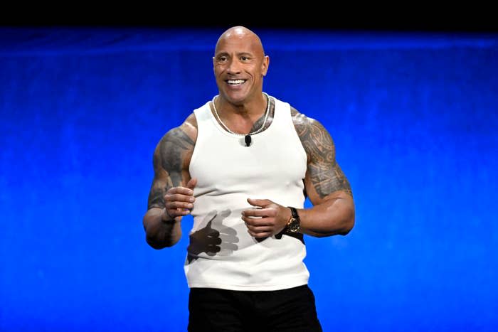 Dwayne Johnson, known as "The Rock," smiles on stage wearing a sleeveless white shirt, showing his tattooed arms, and black pants at a public event