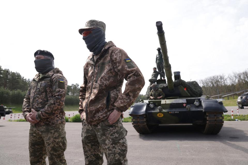 Two members of the tank crew stand in the foreground while other stand on the tank in the background.