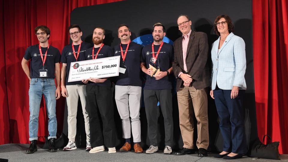  People stand on a stage wearing medals and holding a check. 