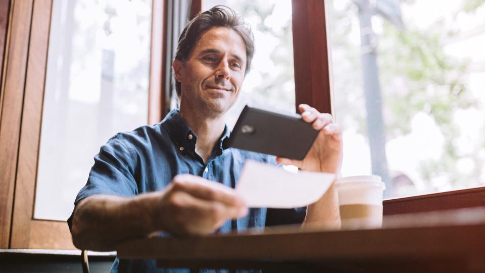 A mature smiling man in his mid 40's takes a picture with his smart phone of a check or paycheck for digital electronic depositing, also known as 