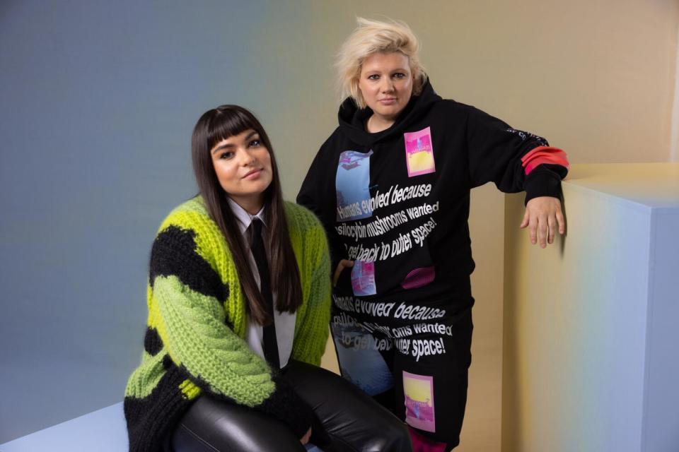 Devery Jacobs and D.W. Waterson pose together while one rests an arm on a tall cube and the other sits on another cube.