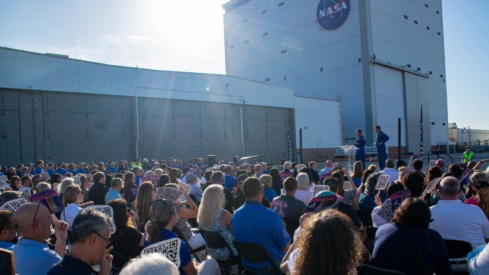 Two men in the distance stand before a tall building blockgin the sun. It has a nasa logo near the top. They address a large crowd, stretching the rest of the frame.