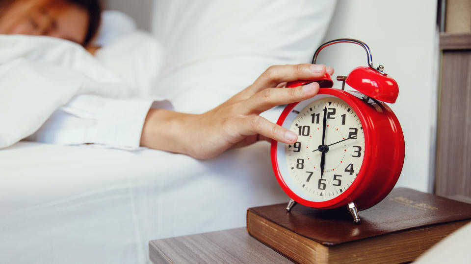 A woman lifts her arm out from under her white duvet in order to turn off her alarm clock