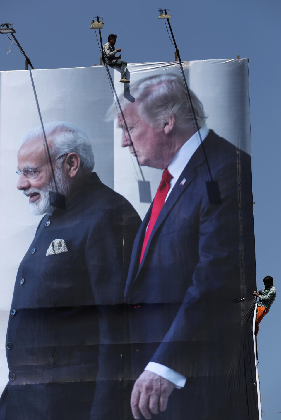FILE - In this Feb. 20, 2020, file photo, workers install a giant hoarding welcoming U.S. President Donald Trump ahead of his visit, in Ahmedabad, India. To welcome Trump, who last year likened Modi to Elvis Presley for his crowd-pulling power at a rally in Houston, the Gujarat government has spent almost $14 million on ads blanketing the city that show the two leaders holding up their hands, flanked by the Indian and U.S. flags. It also scrambled to build a wall to hide a slum from the road Trump and first lady Melania Trump will travel, caught stray dogs, planted exotic trees and is rushing to finish a cricket stadium in time for Trump’s arrival. (AP Photo/Ajit Solanki, File)