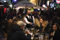 A waiter wears a face mask as people eat and drink outside restaurants in Soho, in London, Wednesday, Nov. 4, 2020. A second lockdown in England is set to come into force on Thursday. It's a big blow to businesses that sweeps away any hopes that the British economy might have recovered by the end of this year a large proportion of the near 25% drop endured in the spring. (AP Photo/Alberto Pezzali)