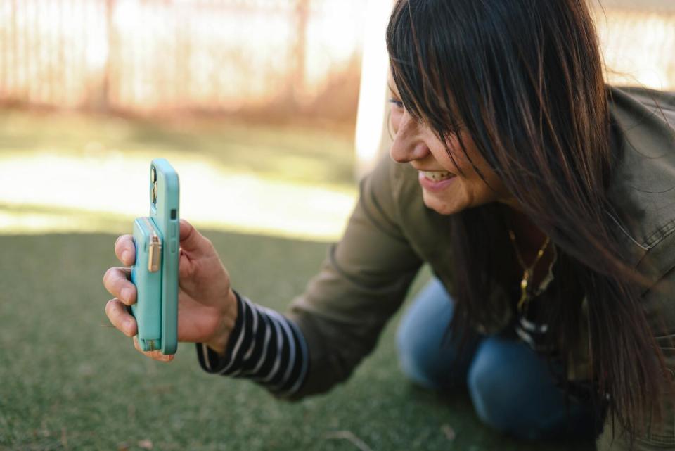 Rita Earl Blackwell holds her teal iPhone as she kneels on the grass to film a dog out of frame.