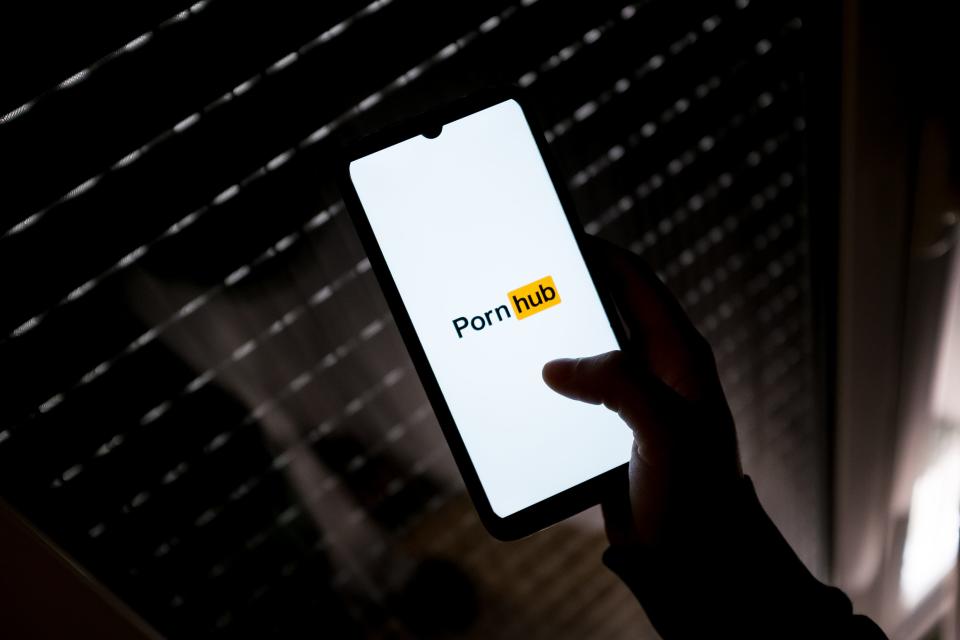 A hand is seen holding a phone, which shows the logo of the company Pornhub.