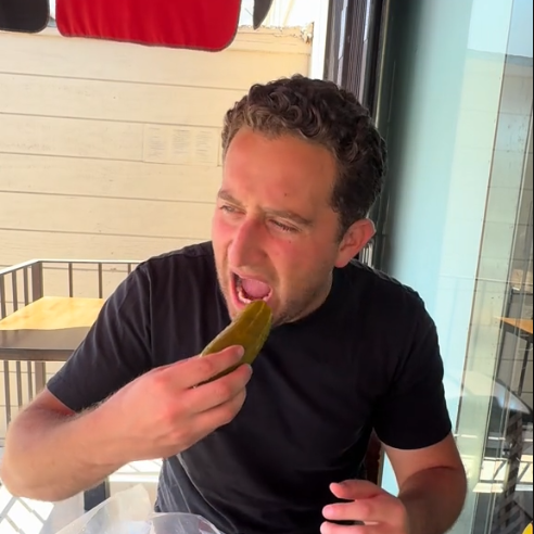 Person in casual clothing eating a pickle at an outdoor table