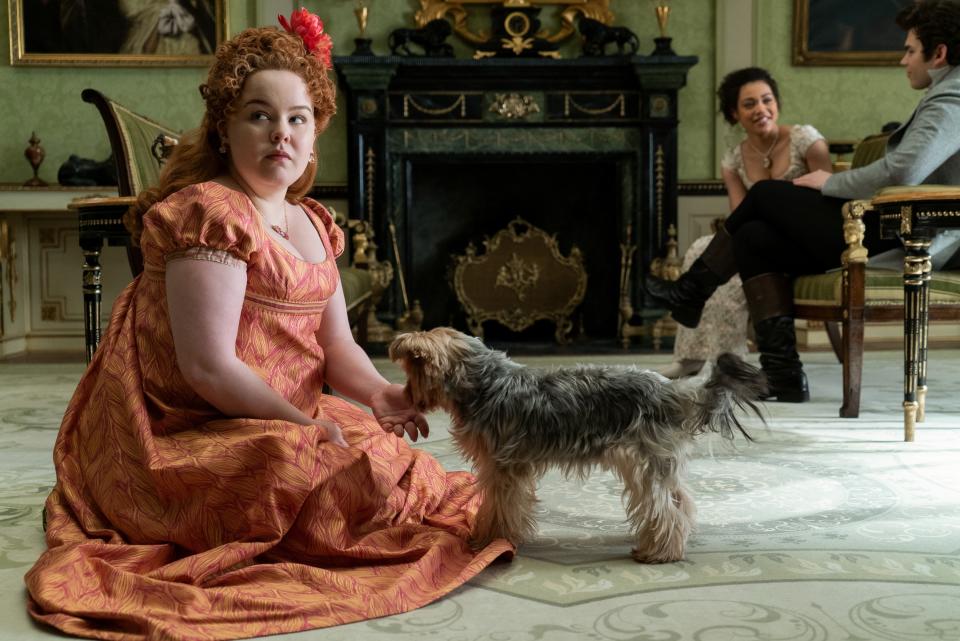 Actress in period costume sitting with dog, two actors in background, in a scene from 'Bridgerton'