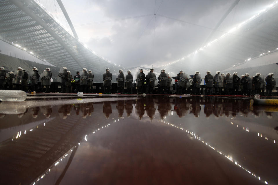 Riot police secure the field as smoke rises from flares in the background during the cup final between Athens's Panathinaikos and Thessalonikis's PAOK at the Olympic Stadium in Athens, Greece Saturday, April 26 2014. More than 300 buses carrying PAOK supporters from the northern Greek city of Thessaloniki were stopped by police at a toll station 34 kilometers (21 miles) north of the capital Athens. The supporters were searched and allowed to proceed, 20 buses at a time, to the Athens Olympic Stadium, where the Greek Cup final between soccer clubs PAOK and Panathinaikos is taking place. Police, fearing clashes between rival fans, directed supporters to different gates of the stadium several hours before the game's start. (AP Photo/Kostas Tsironis)