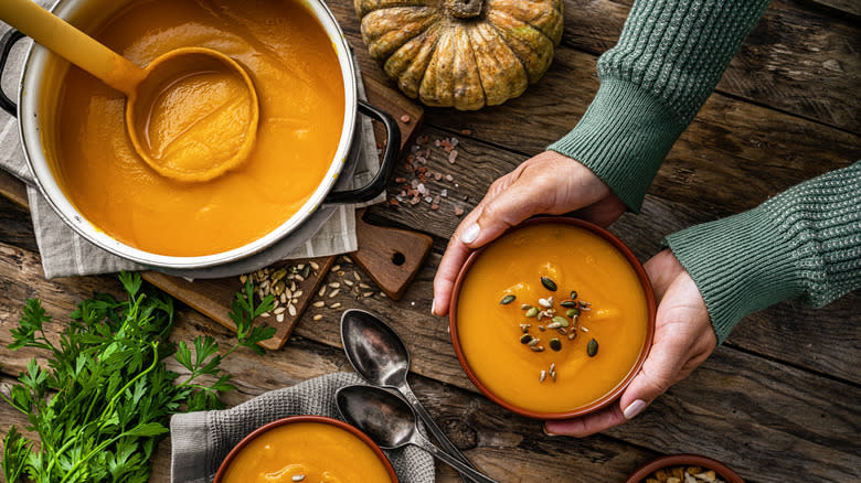 Hands holding bowl of pumpkin soup next to a pot over wooden surface