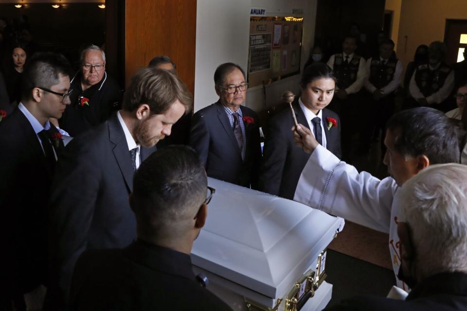 A priest sprinkles holy water on a casket amid a cluster of people, including pallbearers