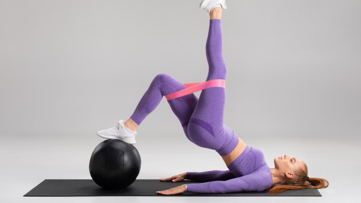  Woman performing a one-leg glute bridge with standing foot on a medicine ball against grey backdrop, wearing a loop band around knees 