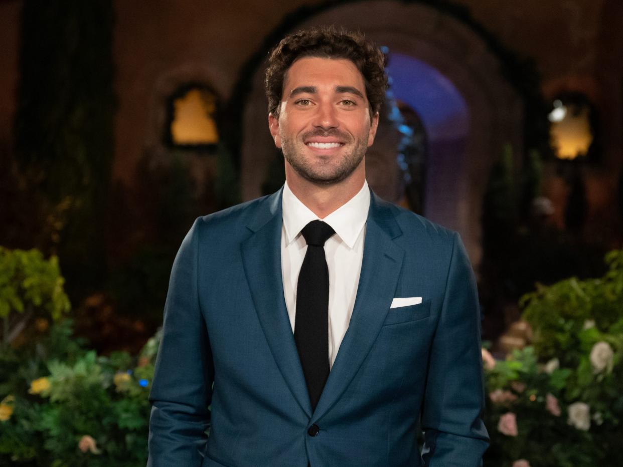 Joey Graziadei smiles in front of the Bachelor mansion in a blue suit, white shirt, and black tie.
