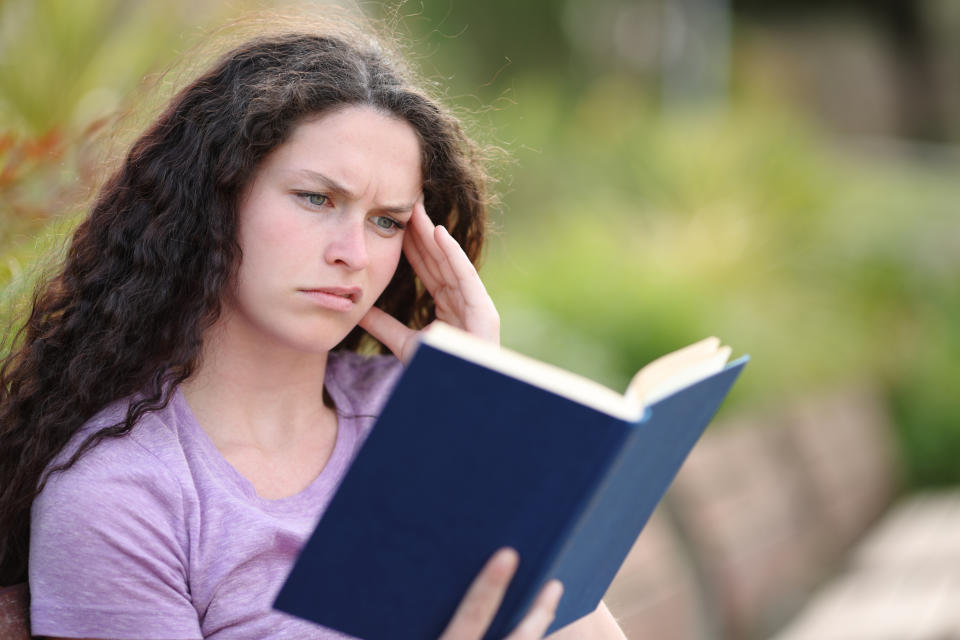 Person with long curly hair reads a book on a bench, looking focused and slightly confused. Wearing a casual shirt