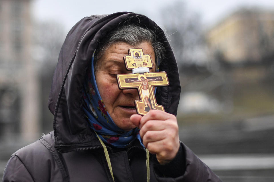 A middle-aged woman in rainwear holds a gold cross painted with Orthodox icons to her forehead.