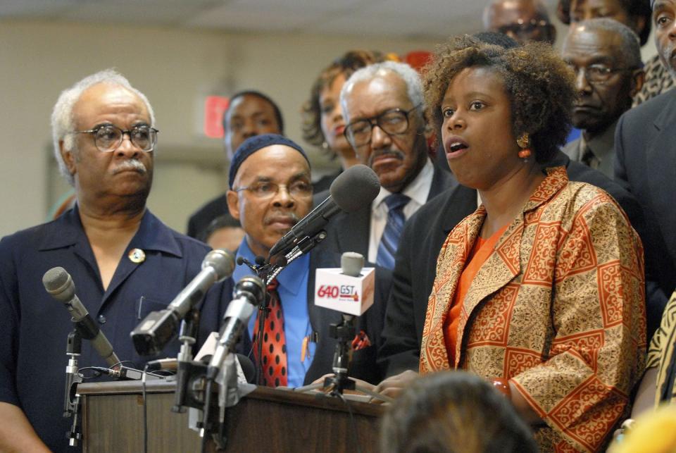 cynthia mckinney stands at a podium with several microphones and speaks, a crowd of people watch from behind her, she wears an orange and gold pattern suit jacket over an orange blouse