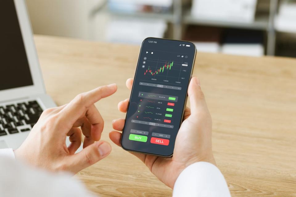 A person is holding a phone with a stock chart on it and Buy and Sell buttons.