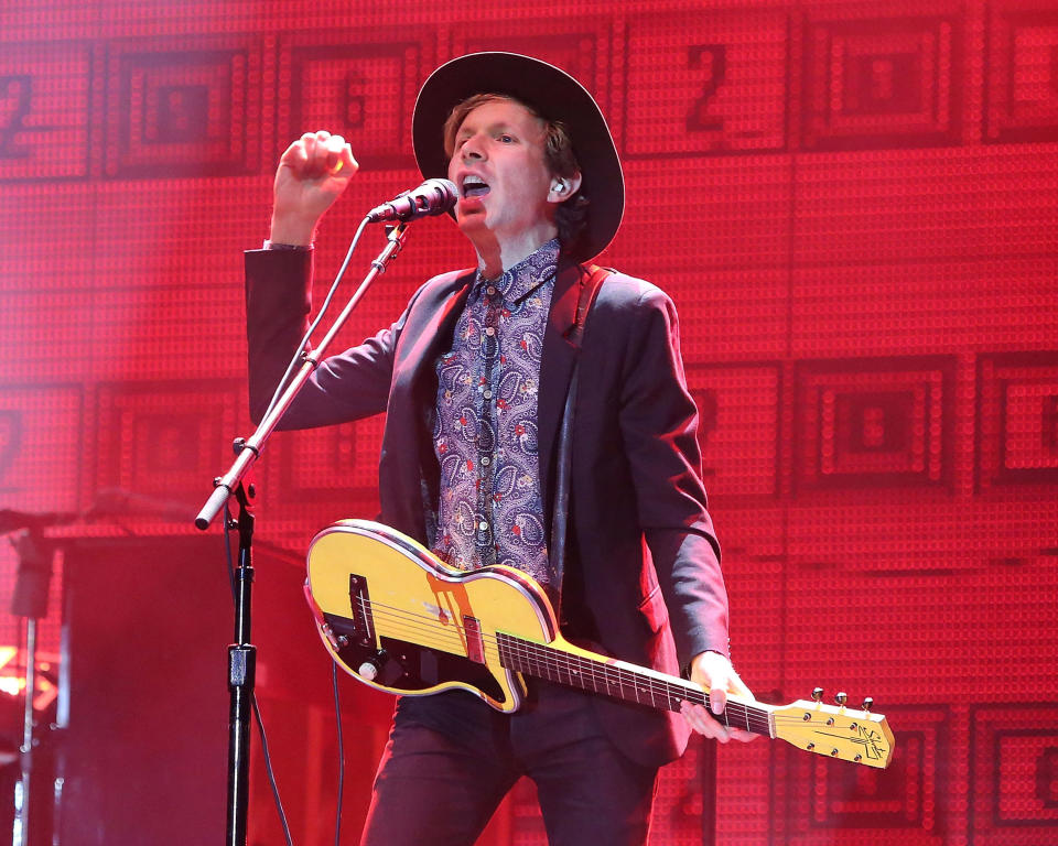 Beck performs on stage wearing a patterned shirt, dark suit, and hat while singing and playing a yellow guitar