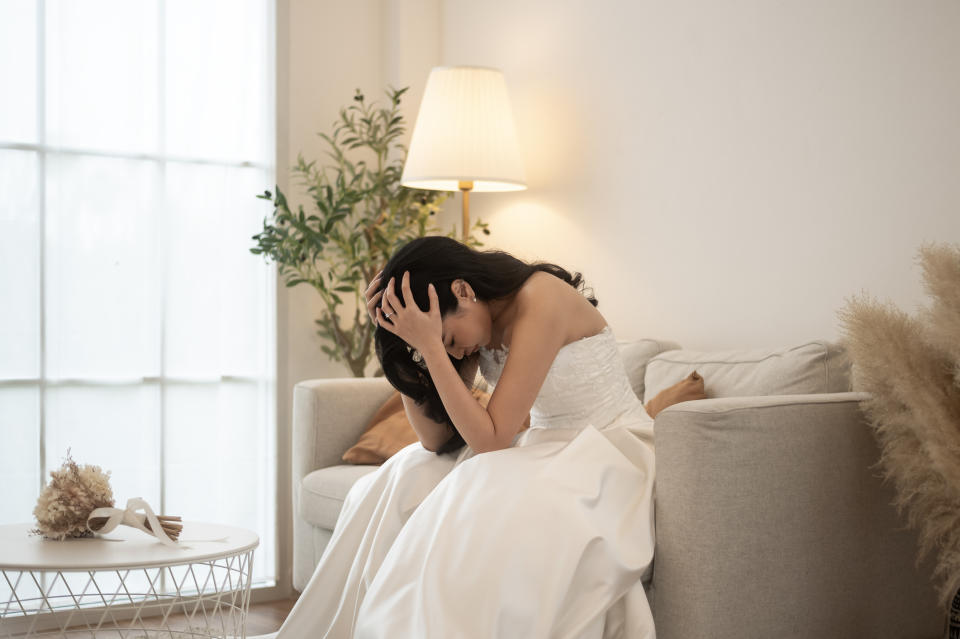 A woman in a wedding dress sits on a beige couch, holding her head in her hands, looking distressed. A lamp and plant are in the background