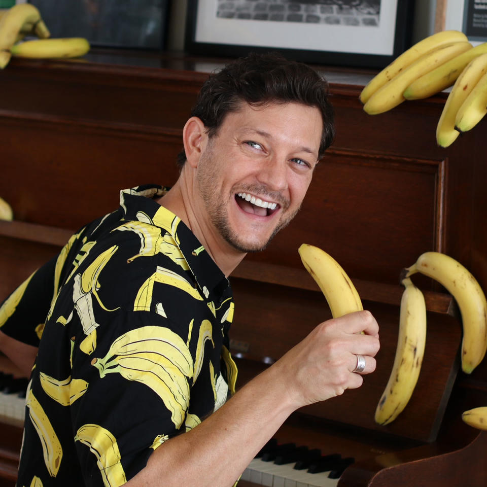 Rob Mills wearing a black shirt with bananas on it, holding a banana and pretending to sing. 