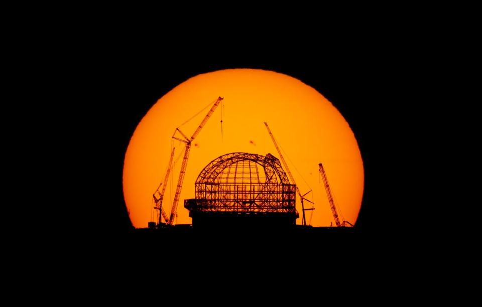 A gigantic view of the sun at sunrise can be seen behind a silhouetted ELT under construction.