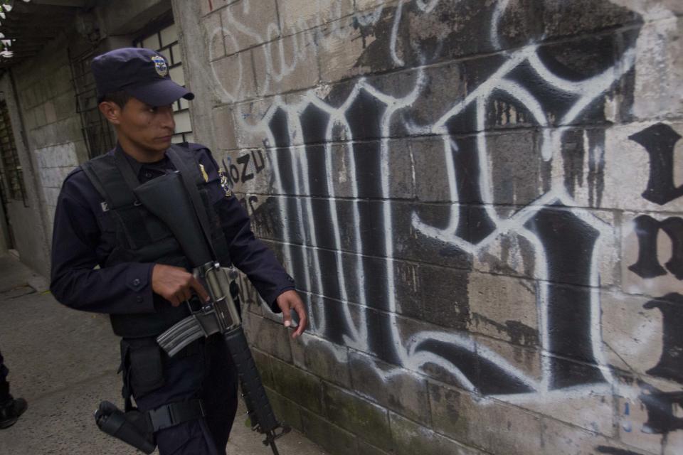 FILE - In this March 7, 2014 file photo, a member of the Salvadoran National Police walks next to symbol of the "Mara Salvatrucha" gang painted on a wall during an anti-gang operative in San Salvador, El Salvador. Public Safety Minister Ricardo Perdomo said the country’s Mara Salvatrucha and Mara 18 street gangs are buying automatic rifles in the black market in Honduras and Guatemala, and also getting them from the brutal Zetas drug cartel. (AP Photo/Esteban Felix, File)