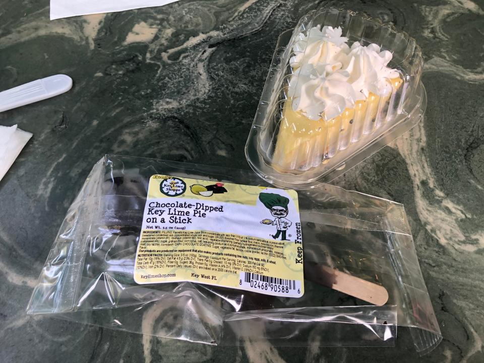 Key-lime pie on a chocolate stick in a plastic wrapping next to a slice of Key-lime pie in a clamshell container
