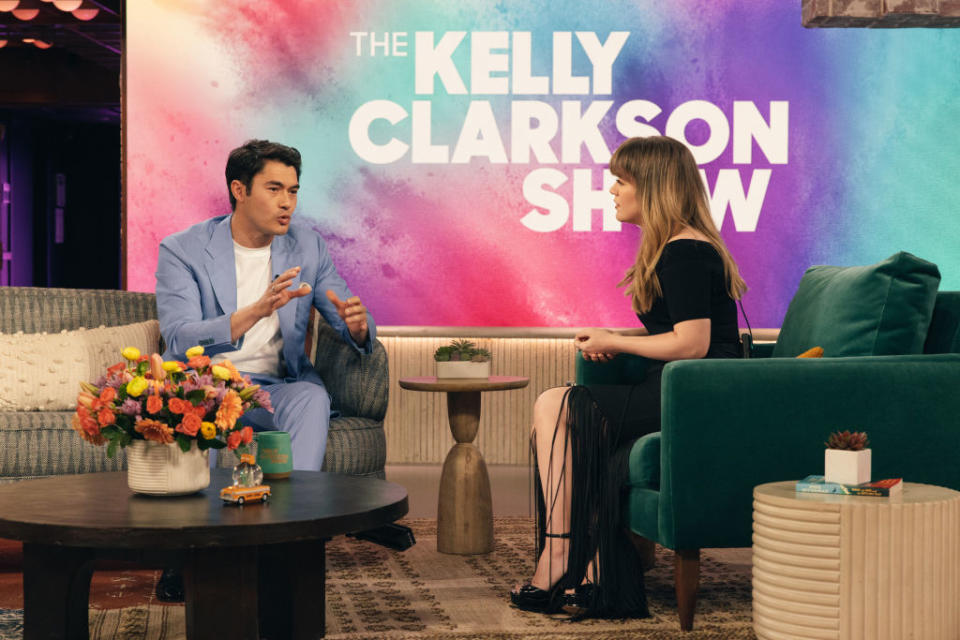 Henry Golding, in a blue suit, chatting with Kelly Clarkson on her show set