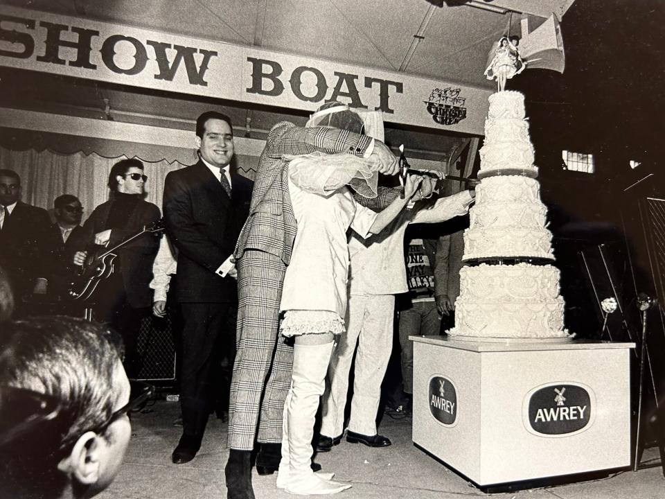 The bridal couple cut the Awrey wedding cake, supervised by a partially hidden baker in a white uniform next to the bride. Famed WKNR deejay Scott Regen grins as Velvet Underground leader Lou Reed, with sunglasses, looks on.