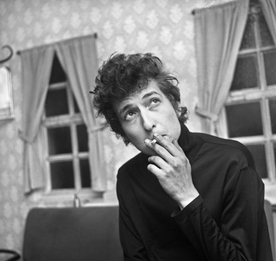 Bob Dylan smoking a cigarette in a room with two curtained windows in the background. He is wearing a dark turtleneck sweater