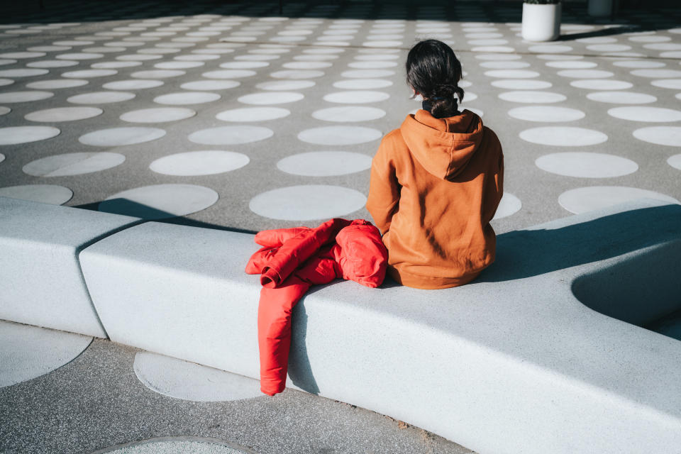 A woman in an orange hoodie sits on a bench in a public square.