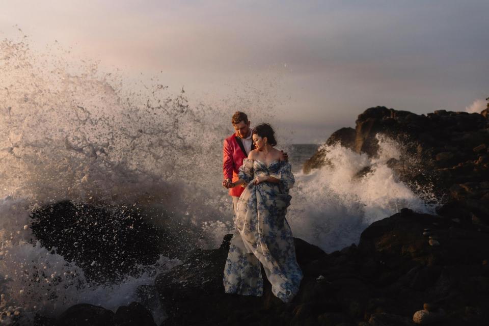 A bride and groom embrace as an ocean wave crests around them.