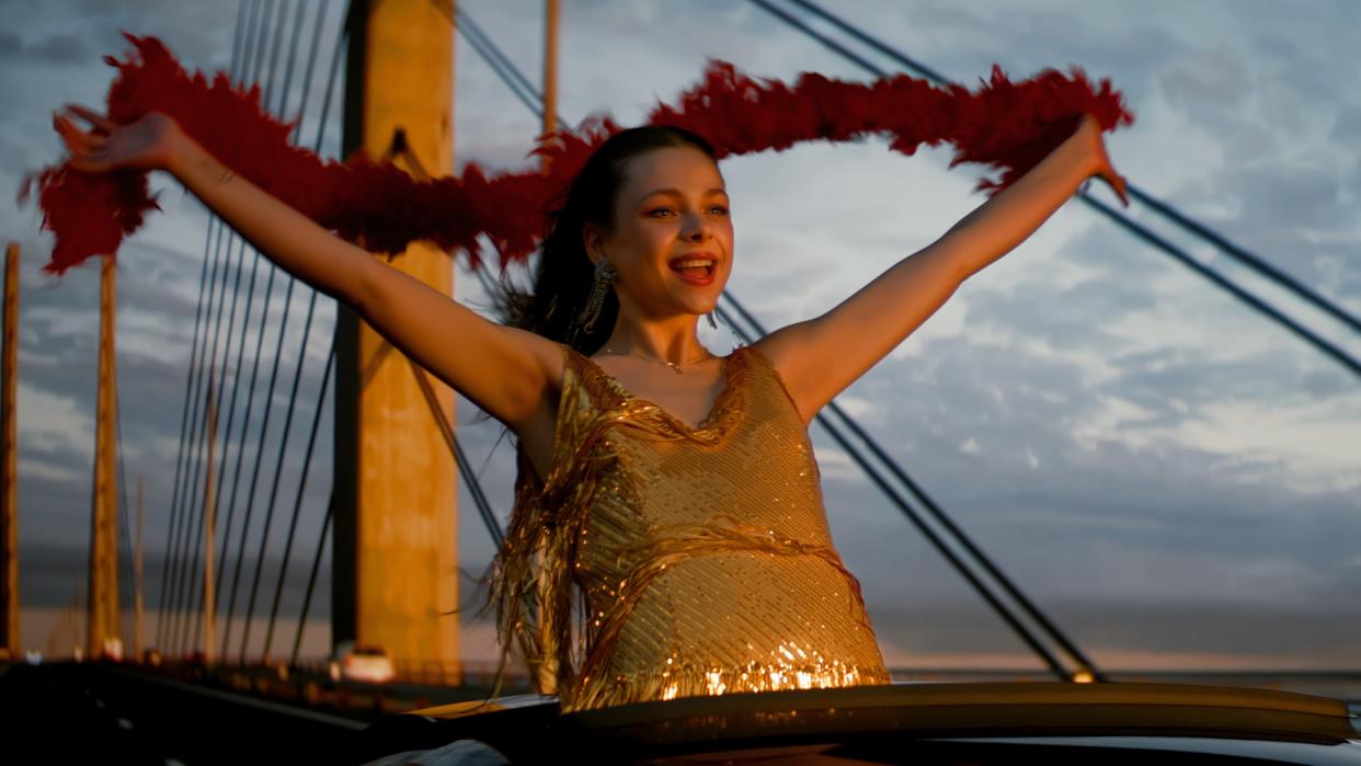  Stella (Alexandra Tyrefors) in a sparkly gold dress and holding a red feather boa out of a sun roof in A Nearly Normal Family episode 1 . 
