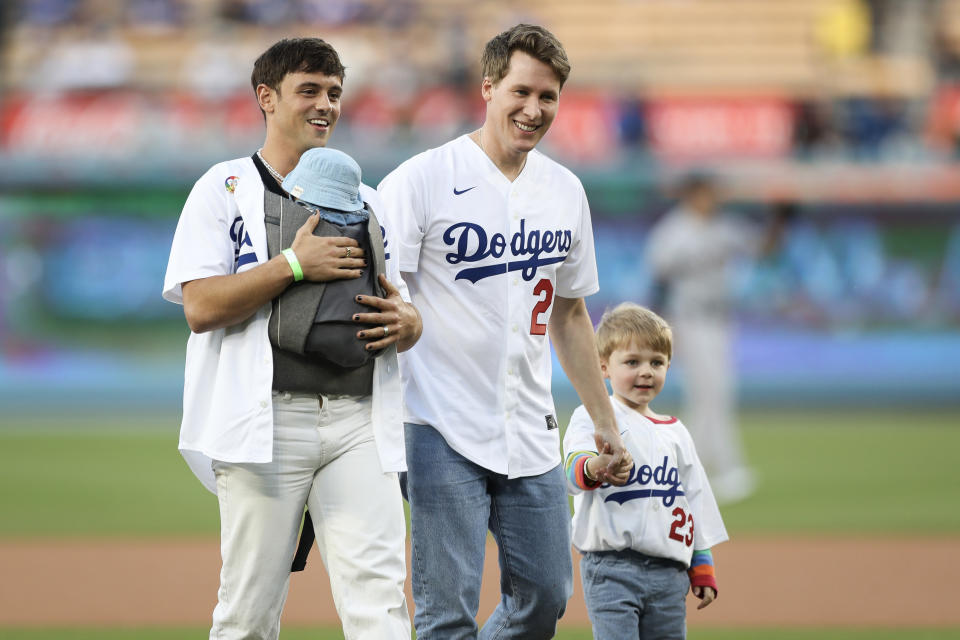 Tom Daley and Dustin Lance Black walk on a baseball field with their child, Robbie, who is wearing a Dodgers jersey and holding their hands, while carrying a baby