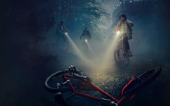 Three of the main characters from Stranger Things, shining their flashlights over a red bicycle on a misty night.