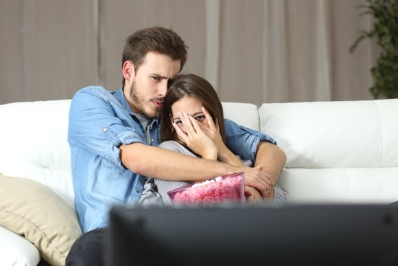 A young couple cuddling on the couch with a bucket of popcorn. The woman peeks at the screen between her fingers.