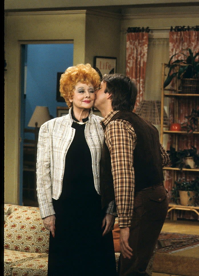 Lucille Ball receives a playful kiss on the cheek from a man wearing a plaid shirt and vest on the set of a 1970s TV show
