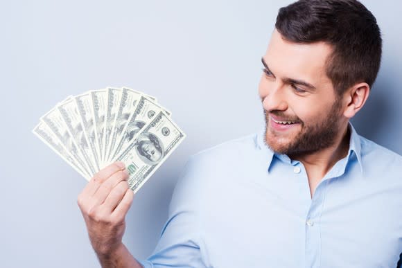 A smiling man holding several 100 dollar bills fanned out in his hand.