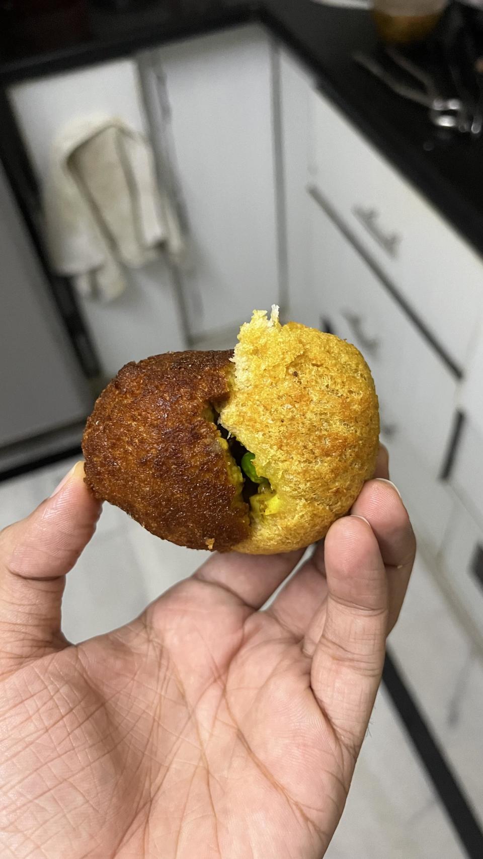 A hand holds a broken stuffed ball of fried dough, revealing a green filling, in a kitchen with white cabinets and a towel hanging on the oven handle