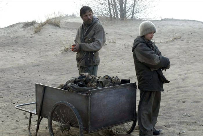 A haggard man and a young boy stand on a beach near a cart with all of their belongings