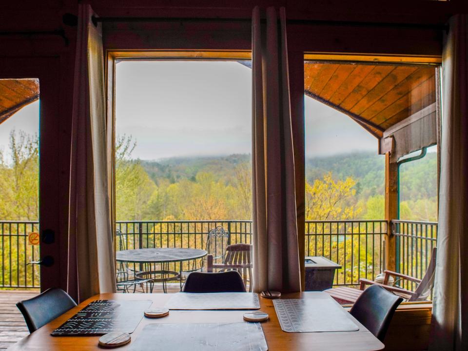 A wood table with four black chairs in front of wide windows showing a patio, trees, and mountains outside on a cloudy day