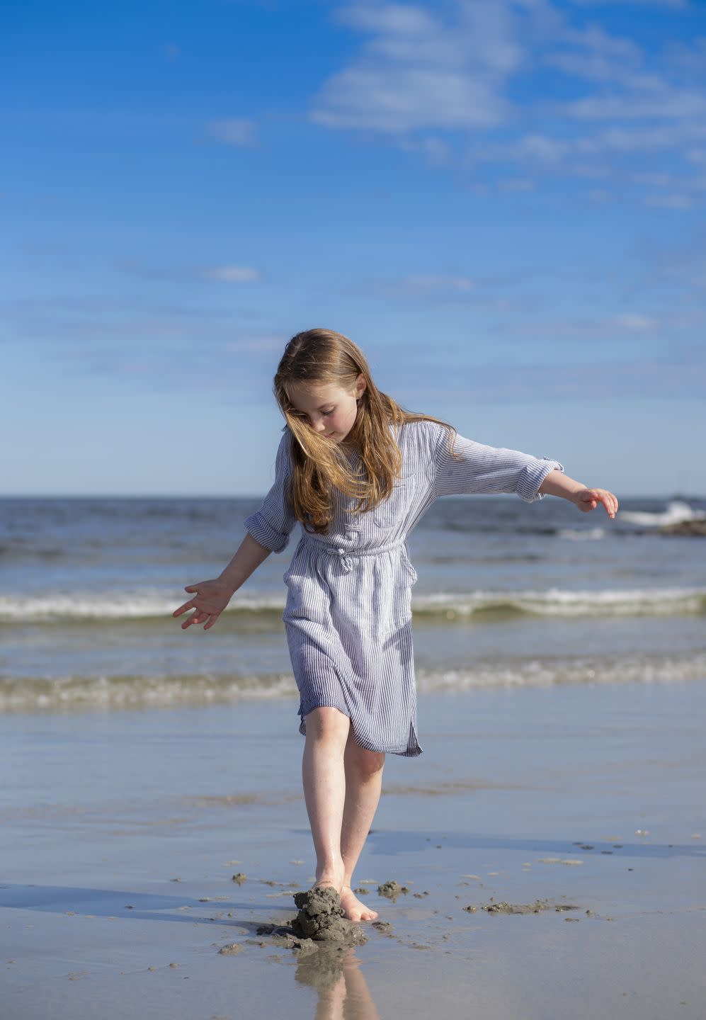 elementary age girl in blue dress walking on a beach and digging feet into the sand