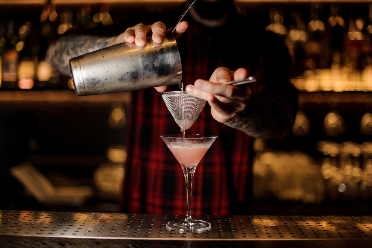 Barman pouring fresh and tasty Cosmopolitan cocktail into an elegant martini glass using shaker and strainer