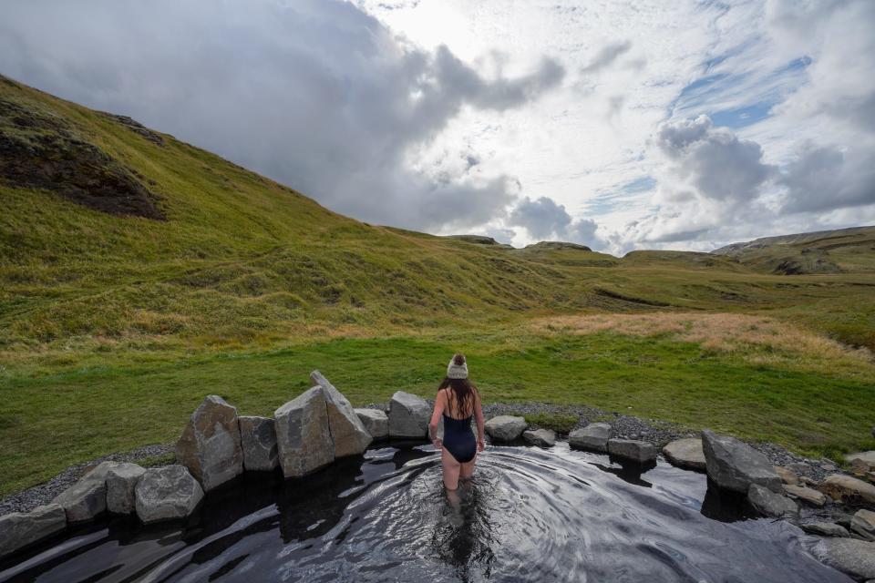 Author Lauren Breedlove standing in a hot spring with a hat on facing the green mountains and skies ahead in Hrunalaug